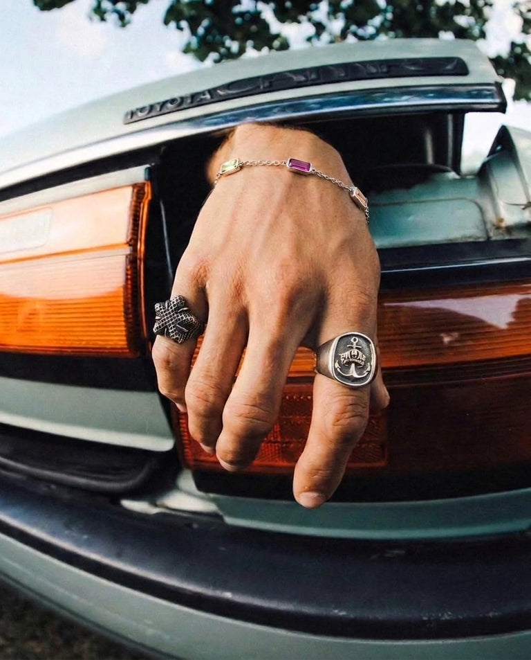 Man's hand wearing a Steelixe stainless steel signet ring and gemstone bracelet, resting on a classic car